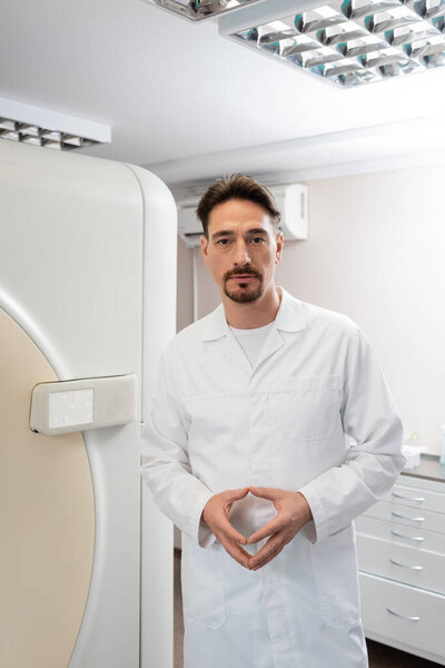 brunette doctor in white coat looking at camera near computed tomography machine in clinic
