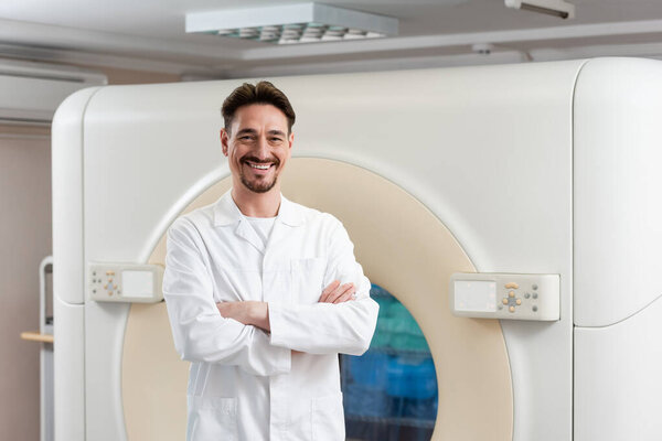 bearded physician in white coat smiling at camera near ct scanner in hospital