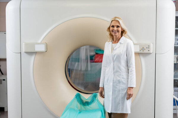 pretty blonde radiologist in white coat smiling at camera near computed tomography machine