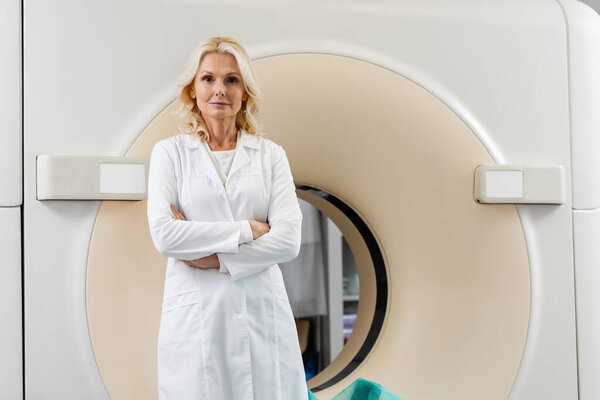 blonde radiologist in white coat standing with crossed arms and looking at camera near ct scanner