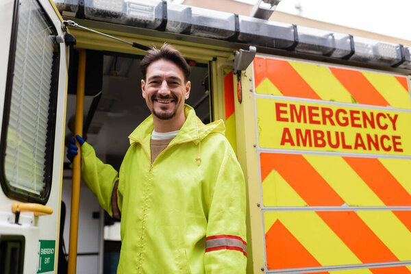 Positive paramedic looking at camera near ambulance car on urban street 