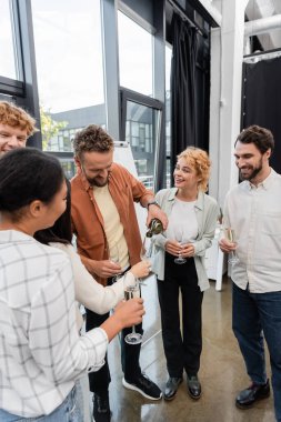 businessman pouring champagne near cheerful interracial colleagues at corporate party clipart