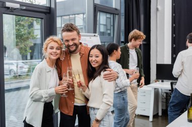 Positive businessman hugging multiethnic colleagues with champagne in office  clipart