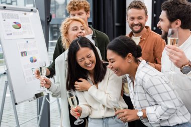 Multicultural business people with champagne laughing near flip chart in office  clipart