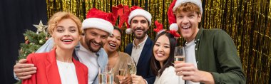 Cheerful interracial business people in santa hats holding champagne near tinsel in office, banner  clipart