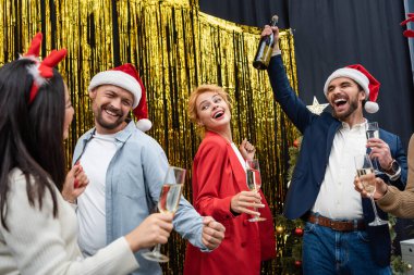 Heyecanlı iş adamı Noel Baba şapkalı, elinde şampanya şişesiyle ofiste dans eden ırklar arası iş arkadaşlarının yanında. 