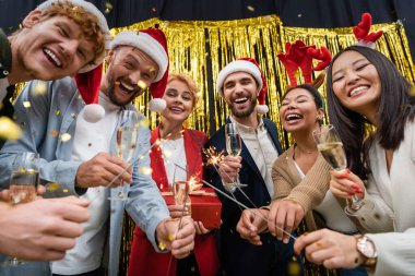 Low angle view of multiethnic business people holding sparklers and champagne during New Year party in office  clipart