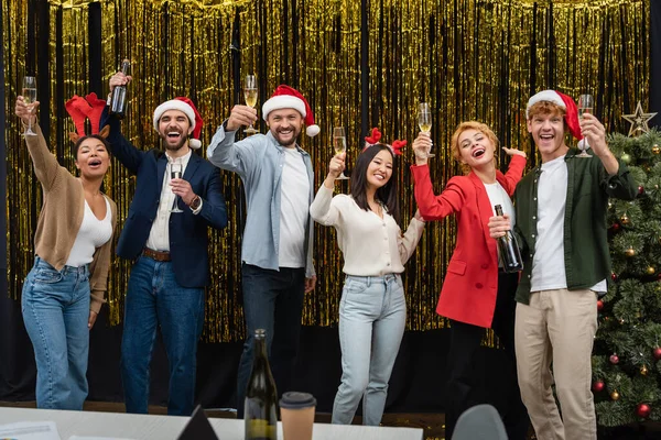 クリスマスツリーの近くにシャンパンを保持サンタ帽子の幸せな多文化ビジネスの人々とオフィスでチンセル — ストック写真