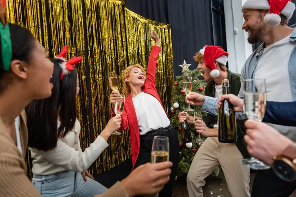 オフィスでシャンパンとクリスマスツリーと多民族の同僚の近くで踊るビジネスマン女性 — ストック写真