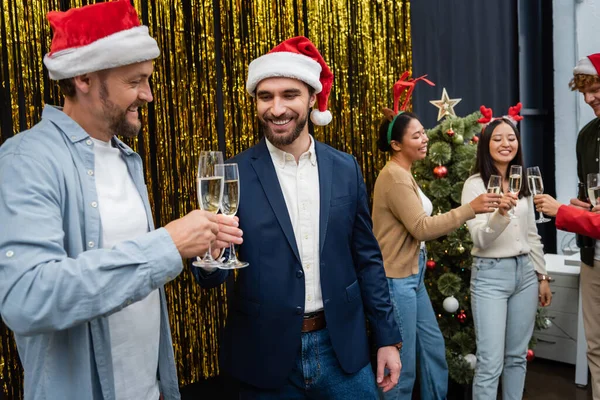 オフィスで多民族の同僚やクリスマスツリーの近くにシャンパンをクリックサンタ帽子の笑顔ビジネスマン — ストック写真