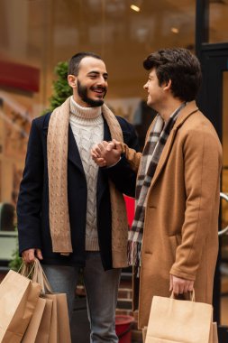 pleased and stylish gay partners with shopping bags holding hands while looking at each other near blurred showcase clipart