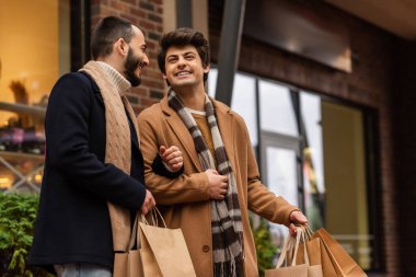 trendy gay men in scarfs holding shopping bags and smiling on city street clipart