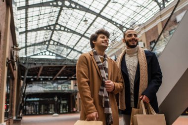 Moda giysiler içinde neşeli eşcinsel erkek arkadaşlar şeffaf çatı altında alışveriş torbalarıyla caddede duruyorlar.