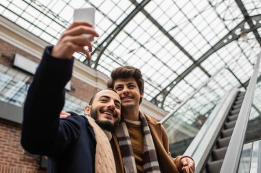 Sakallı eşcinsel adam, yürüyen merdivende genç erkek arkadaşıyla selfie çekiyor.
