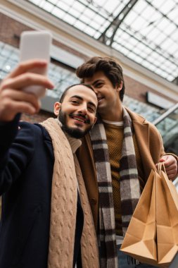 Neşeli sakallı adam erkek arkadaşıyla dışarıda alışveriş torbaları tutarken selfie çekiyor.