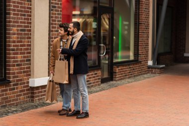 full length of bearded gay man with shopping bags pointing at showcase near young boyfriend clipart