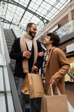 Alışveriş torbaları ve kağıt bardaklarıyla yürüyen merdivende birbirlerine gülümseyen şık eşcinsel çift.