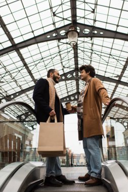 Alışveriş torbaları ve kağıt bardaklarla gülümseyen eşcinseller şeffaf çatı altında yürüyen merdivenin üstünde duruyorlar.