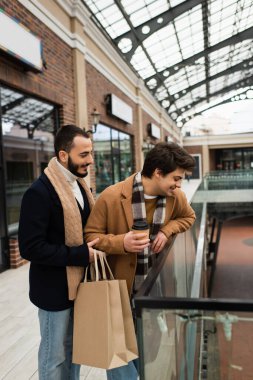 gay man with paper cup looking down near glass fence and bearded boyfriend with shopping bags clipart