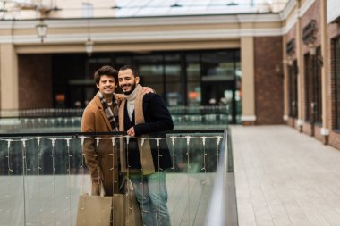 Moda ve mutlu gay adamlar alışveriş torbaları ile cam çitlerin yanında duruyor ve bulanık bina