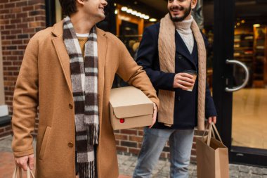 partial view of stylish and happy gay men with purchases and coffee to go near blurred showcase on urban street clipart