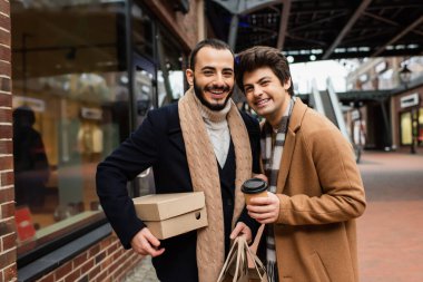 happy and fashionable gay couple with purchases and coffee to go looking at camera near showcase on street clipart
