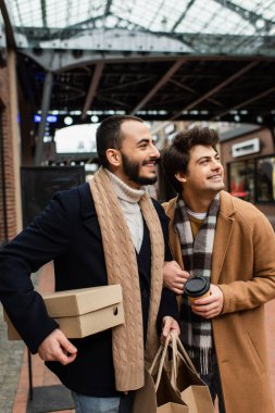 trendy gay couple with purchases and coffee to go smiling and looking away on blurred street clipart