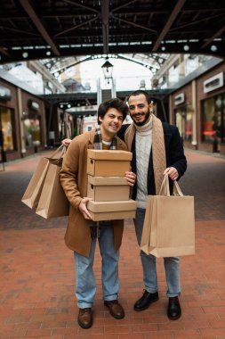 full length of joyful homosexual men with shopping bags and shoeboxes near building with shops on background clipart