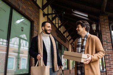 cheerful man with shoebox looking at bearded boyfriend holding shopping bags near building with showcases clipart