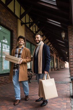 full length of happy gay couple in trendy clothes standing with purchases near building with stores clipart