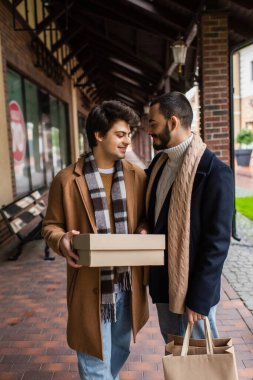 young gay man in checkered scarf holding shoebox near smiling boyfriend with shopping bags clipart