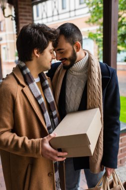 young gay man with closed eyes holding shoebox while standing face to face with happy bearded boyfriend  clipart