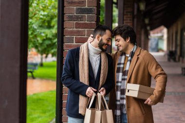 pleased and fashionable gay couple with purchases standing with closed eyes near brick column clipart