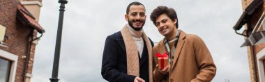 young and happy gay men looking at camera near small gift box on city street, banner clipart