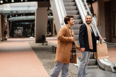 cheerful gay couple in fashionable outfit holding hands while standing with shopping bags near escalator clipart