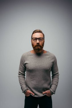 Şık sakallı, kazaklı, gri pantolonun cebinde el ele tutuşan adam. 