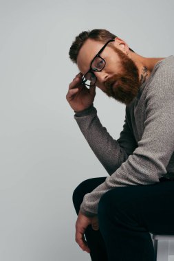 Sakallı ve dövmeli bir adam gri sandalyede izole bir şekilde otururken kameraya bakıyor. 