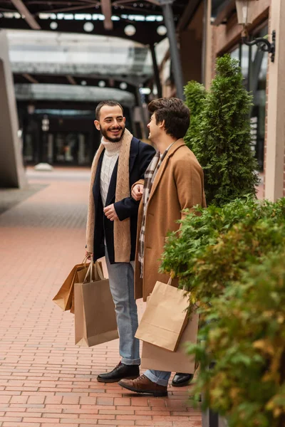 时尚的同性恋伴侣 购物袋在街上的绿色植物旁互相微笑 — 图库照片