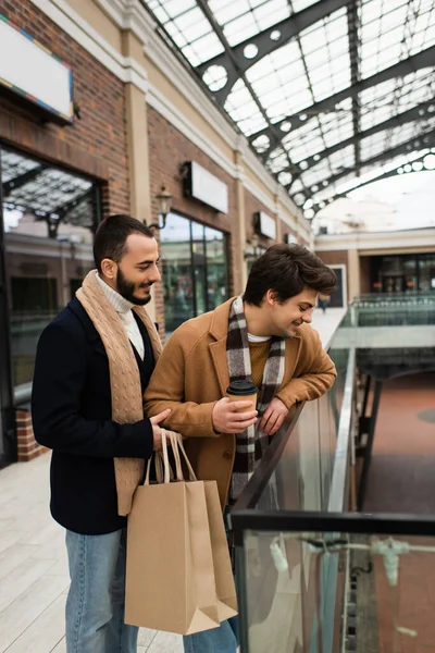 gay man with paper cup looking down near glass fence and bearded boyfriend with shopping bags