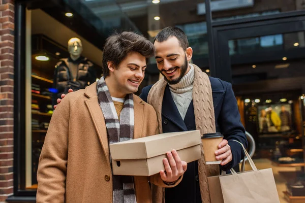 笑容满面的男人拿着咖啡去看鞋盒 男朋友手里拿着鞋盒在街上模糊的橱窗旁 — 图库照片