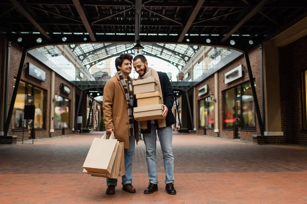 都会の通りに店があるビルの近くにショッピングバッグや靴箱を持つ流行のゲイカップルの完全な長さ — ストック写真