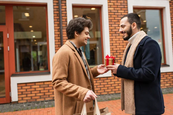 買い物袋を持った若いゲイの男のサイドビュークリスマスプレゼントを路上の店の近くのひげを生やしたボーイフレンドに — ストック写真