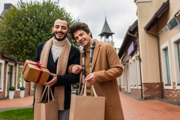 stock image young gay partners in trendy clothes holding shopping bags and Christmas present while smiling at camera outdoors