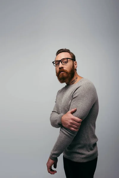 Stylish bearded man in sweater and eyeglasses looking at camera isolated on grey