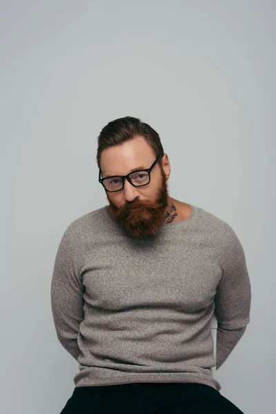 Portrait of stylish tattooed man in eyeglasses looking at camera while sitting on chair isolated on grey