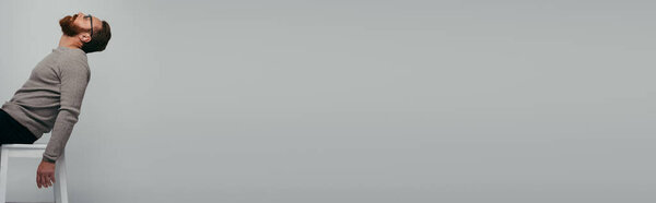 Side view of bearded man in eyeglasses sitting on chair isolated on grey, banner 