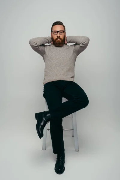 stock image Full length of stylish man in eyeglasses touching neck and looking at camera while sitting on chair on grey background 