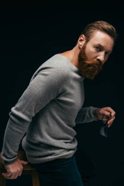Süveter giyen, gözlüklü ve siyah sandalyenin yanında duran şık bir adam. 