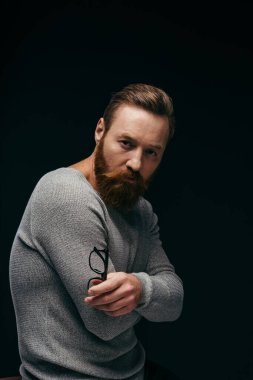 Gri kazaklı şık bir adam siyah kollarıyla çapraz kollar tutarken gözlük tutuyor. 