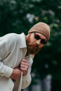 bearded man in stylish sunglasses and beanie hat standing in white shirt jacket posing near green leaves clipart